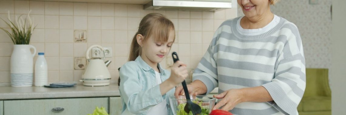Little girl is cooking salad mixing vegetables in bowl and chatting to cheerful granny in kitchen at home. Modern lifestyle, meals preparation and family concept.